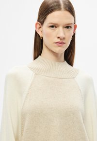 Μπεζ πλεκτό πουλόβερ με ψηλό, ριμπ πλεκτό γιακά και μανίκια ραγκλάν. Απαλή υφή, μετάβαση από ανοιχτό μπεζ σε εκρού στα μανίκια.