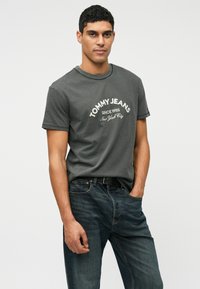 Γκρι βαμβακερό T-shirt με την ένδειξη "TOMMY JEANS" τυπωμένη σε λευκό, συνδυασμένο με σκούρα μπλε τζιν παντελόνι. Κοντά μανίκια και στρογγυλή λαιμόκοψη.