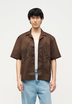 Jeune homme portant une chemise marron à manches courtes boutonnée sur un débardeur blanc et un jean bleu clair, debout devant un fond uni.