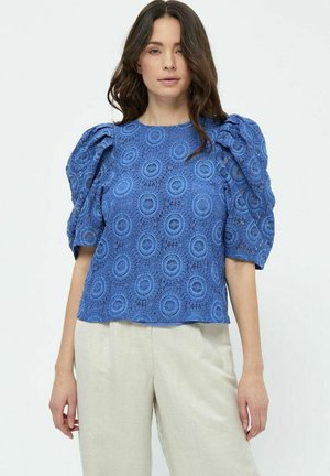 Blaues Spitzenblusenshirt mit kreisförmigen Mustern, Puffärmeln und rundem Halsausschnitt, kombiniert mit hellen Hosen.