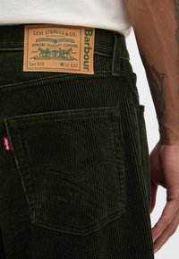 Πράσινο παντελόνι κοτλέ με δερμάτινη ετικέτα που αναγράφει «Levi Strauss & Co.» και «Barbour». Ορατές ραφές και λεπτομέρεια πλαϊνών τσεπών.