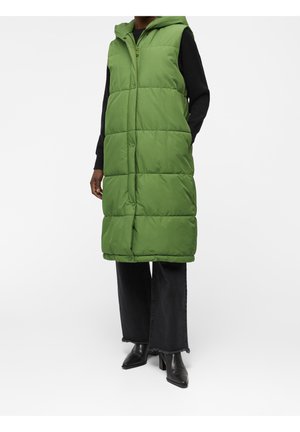 Personne portant une longue doudoune sans manches verte avec une capuche, un pantalon noir à jambes larges, une chemise noire et des bottines noires à talons.