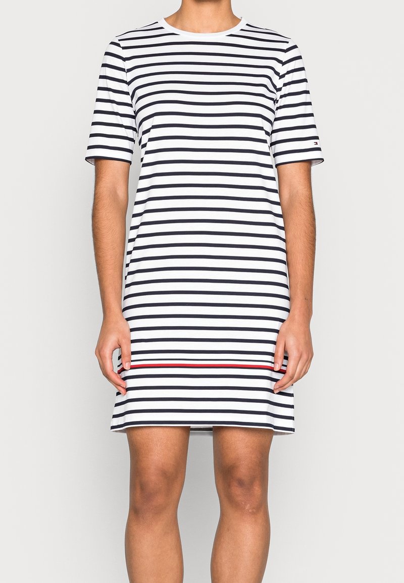 Personne portant une robe blanche courte à manches courtes, longueur genoux, avec des rayures noires horizontales et une seule rayure rouge près de l’ourlet.