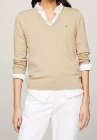 Beżowy sweter z dekoltem w serek, wykonany z miękkiego materiału, z prążkowanymi mankietami i dołem, noszony na białej koszuli z kołnierzykiem. Mały detal z logo na klatce piersiowej.