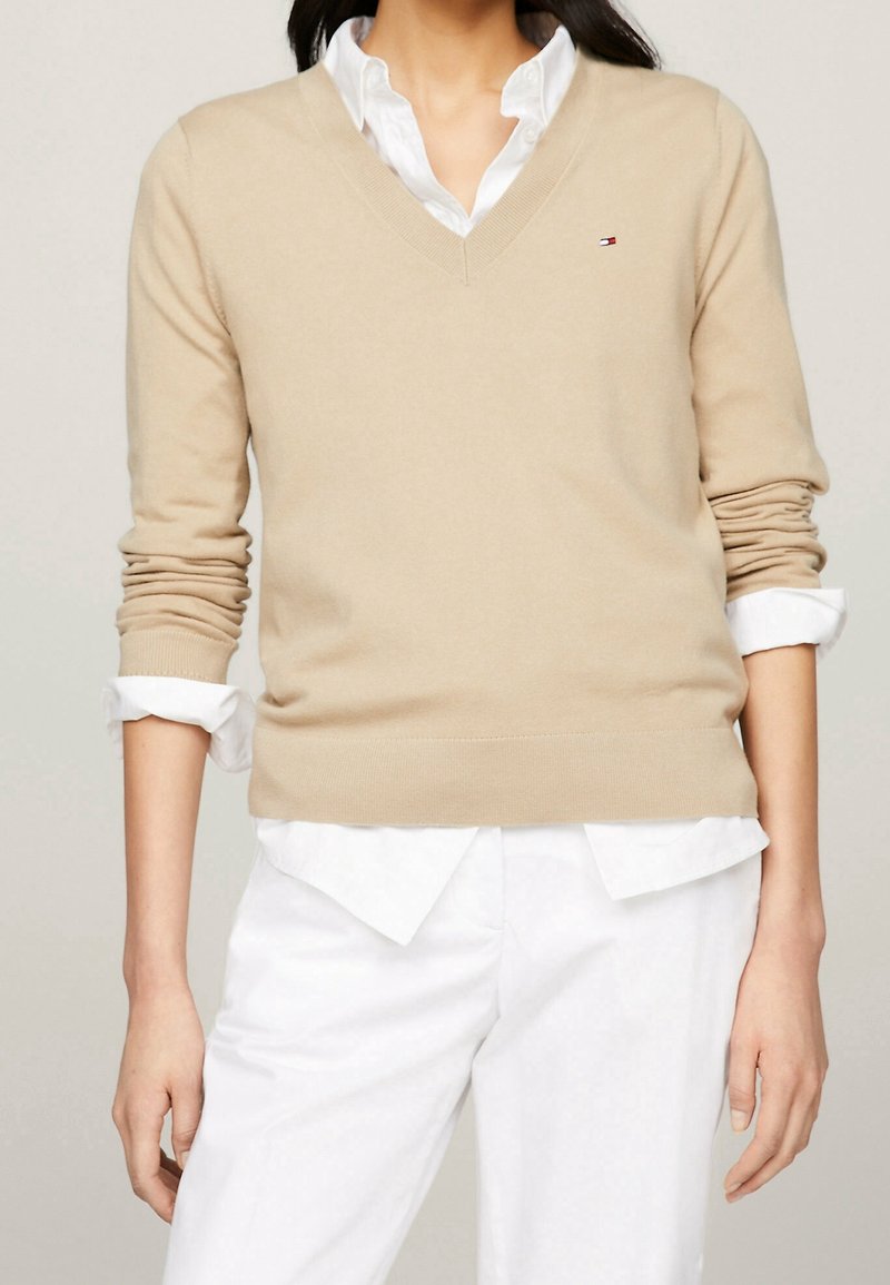 Beżowy sweter z dekoltem w serek, wykonany z miękkiego materiału, z prążkowanymi mankietami i dołem, noszony na białej koszuli z kołnierzykiem. Mały detal z logo na klatce piersiowej.