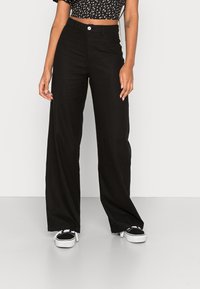 Zwarte wijde broek van stof met een gladde textuur. Kenmerkt zich door een hoge taille en een knoopsluiting aan de voorkant. Draagt met zwarte sneakers.