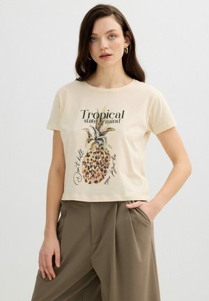 Mujer con cabello largo y castaño que lleva pendientes de aro dorados, una camiseta beige con una piña y texto, y pantalones amplios de pierna ancha de color verde oliva.