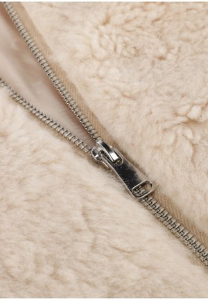 Beige fleece stof met een gladde zilveren rits. Het materiaal heeft een gestructureerd, pluche oppervlak met kleine bobbels.