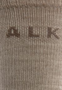 Chaussette en tricot beige avec une texture lisse, présentant les lettres "A L K" dans une couleur brun légèrement plus foncé sur le côté.