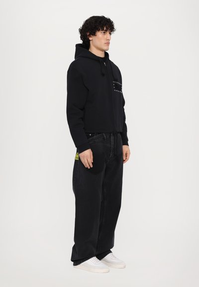 Veste à capuche zippée noire avec une poche texturée, associée à un jean noir oversize et des baskets blanches. Étiquette jaune notable sur le jean.