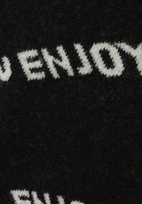 Svart stickad tyg med vit text som säger "DU NJUTER", med en mjuk, fluffig textur och ett grovt vävmönster.