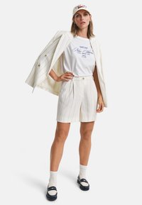 Witte streepjesblazer, wit T-shirt met blauwe tekst, bijpassende shorts, witte sokken en marineblauwe instapper. Pet met rood-wit logo.