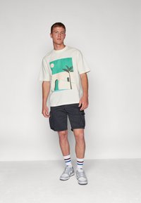 Light cream t-shirt with a teal and beige graphic featuring a sun, palm tree, and abstract landscape; paired with dark shorts and gray sneakers.