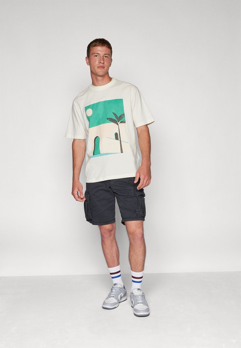 Camiseta de crema claro con un gráfico en teal y beige que presenta un sol, una palmera y un paisaje abstracto; combinada con pantalones cortos oscuros y zapatillas grises.