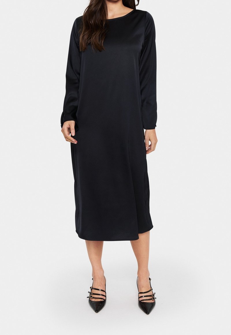 Femme portant une longue robe noire en satin à manches longues et des talons noirs pointus avec trois brides et boucles.