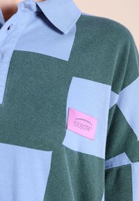 Polo rayé horizontal vert et bleu avec une texture douce et un patch logo Oxbow rose sur le côté gauche. Présente un col classique.