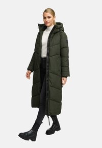 Žena oblečená v dlhom olivovom zelenom páperovom kabáte s kapucňou, bielom roláku, čiernych úzkych džínsach a čiernych hrubých čižmách kráča vpred.