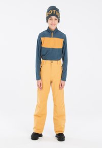 Fleece pullover met een blauwe bovenkant en oranje streep; gele skibroek; zwarte schoenen; gebreide muts met "PROTES" in gele letters.