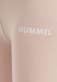Jasnoróżowe legginsy wykonane z miękkiego, elastycznego materiału, z białym logo "HUMMEL" i napisem "SPORTSWEAR SINCE 1923" w pobli�żu talii.