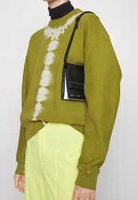 Zielony sweter tie-dye z opadającymi ramionami, w zestawieniu z żółtymi spodniami z wysokim stanem. Czarna torba na ramię z białymi akcentami.