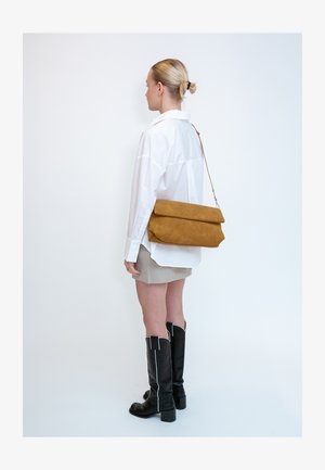 Žena s blond vlasmi v nízkom drdole nosí bielu oversize košeľu, béžovú sukňu, čierne čižmy po kolená a nesie veľkú hnedú kabelku cez rameno.