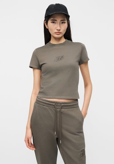 Νεαρή γυναίκα που φοράει ένα καφέ τοπ κοντό t-shirt και αντίστοιχο παντελόνι φόρμας με ρυθμιζόμενο κορδόνι, με μαύρο καπέλο, στέκεται μπροστά σε ένα απλό φόντο.