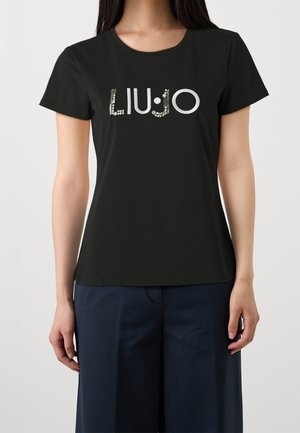 Kobieta w czarnej koszulce z krótkim rękawem z napisem "LIU·JO" wykonanym z srebrnych ćwieków, ubrana w niebieskie szerokie spodnie, stojąca na jednolitym tle.