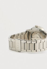 Brățară de ceas din metal argintiu cu închidere și fața de ceas rotundă parțial vizibilă, care arată părțile interne mecanice.