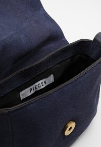 Námořnická semišová kabelka s černým podšívkou. Obsahuje viditelnou etiketu "PIECES" a kulaté zlaté zapínání na patent.