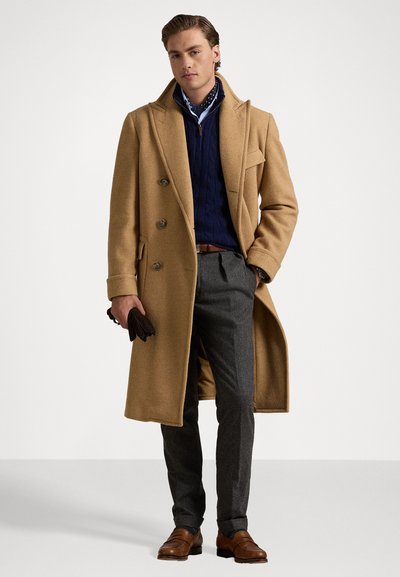 Beige wollen overcoat met dubbele knoopsluiting, marineblauwe kabelgebreide trui, grijze broek en bruine loafers. Zwarte handschoenen in één hand gehouden.