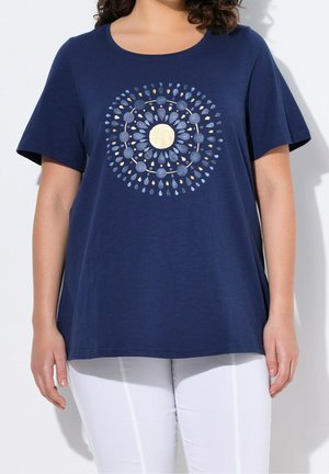Woman wearing a navy blue T-shirt with a circular pattern of gold and blue shapes, paired with white pants against a white background.