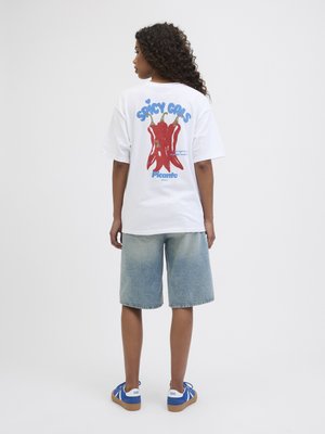 Hvit oversized t-skjorte med grafikk av rød chilipepper og blå tekst "SPICY GALS" på baksiden. Lyst blå denimshorts og blå joggesko.
