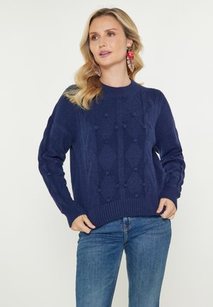 Vrouw draagt een marineblauwe gestructureerde gebreide trui en blauwe jeans, met golvend blond haar en rode bloemige oorbellen, staand tegen een witte achtergrond.