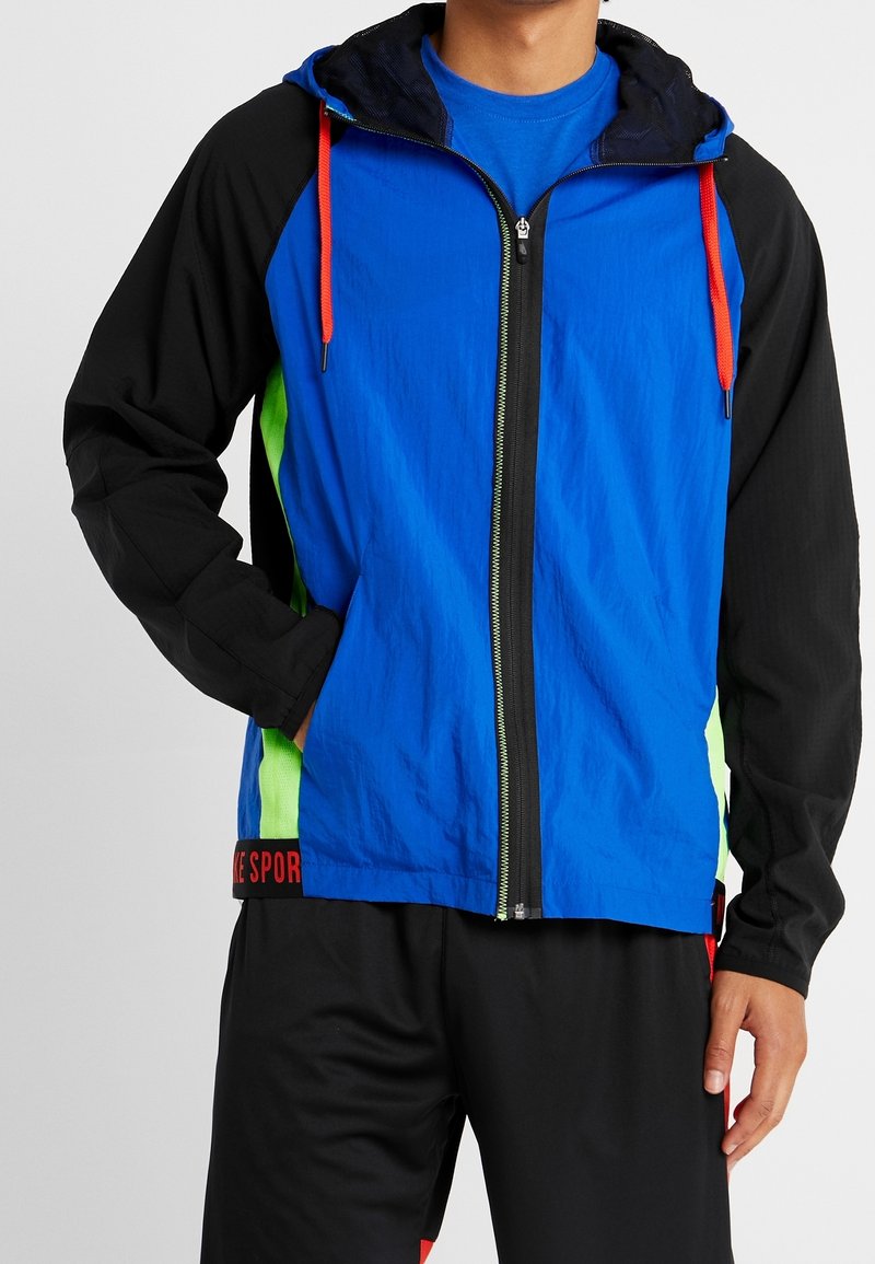 Man som bär en blå zip-up-jacka med svarta ärmar, neongröna sidopaneler, röda dragskor och svarta byxor med röda accenter.