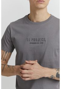 Grå bomull t-skjorte med rund hals, med diskret brodert tekst "11 PROSJEKT. DANMARK ETABLERT. 2020" på brystet.