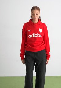 Crvena dugoruka polo majica s ovratnikom, s bijelim "adidas" logom i grbom. Uparena s crnim širokim hlačama viskog struka.