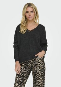 Tamno sivi, pleteni pulover s V-izrezom i čipkastim rubom, uparen s hlačama širokih nogavica u leopard uzorku. Mekana tkanina, ležeran kroj, vidljive džepove.