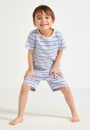 Smilende ung dreng står med benene fra hinanden og hænderne hvilende på hans blå og hvide stribede korte pyjamas mod en ensfarvet baggrund.