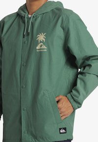 Grøn letvægtsjakke med hætte, der har et palmetræ- og bølgelogo i beige, trykknapper og elastiske manchetter. Inkluderer sidelommer.