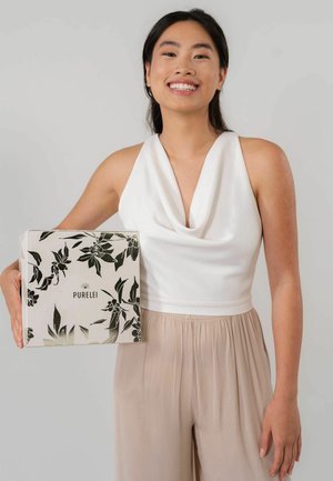 Weißes Oberteil mit Kapuze, kombiniert mit beigen, plissierten Hosen. Das Model hält eine quadratische Box mit floralem Design in Schwarz und Gold, auf der der Text "PURELEI" steht.