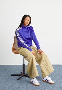 Lila zipjacka med dragkedja och vita ränder, beige vida cargobyxor med fickor som har dragkedjor, och vita sneakers med lila detaljer.