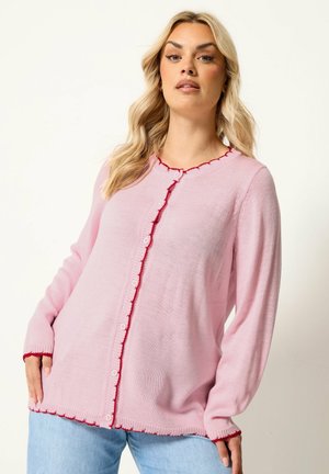 Blond kvinde iført en lyserød knapcardigan med rød kant og lyseblå jeans, stående mod en ensfarvet hvid baggrund.