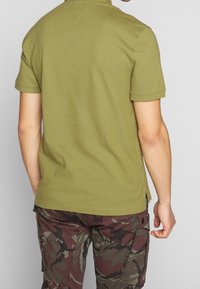 Olivgrön kortärmad polo med krage och sidslitsar, gjord av texturerat tyg. Bärs med burgundy camouflaged byxor.