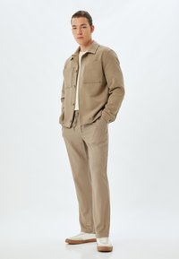 Beige shirt jacket met twee grote borstzakken, gecombineerd met bijpassende broeken en witte sneakers met een tan loopzool. Over het geheel een gladde textuur.