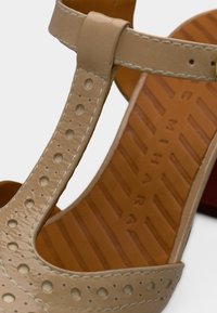 Sandale en cuir beige avec dessus perforé, bride de cheville et semelle intérieure marron texturée. Présente des détails de couture et un petit talon.