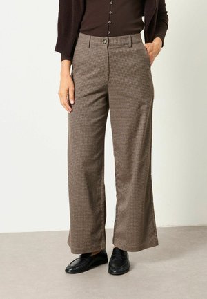 Femme portant un pantalon large à carreaux marron, un haut noir boutonné, un cardigan foncé et des mocassins noirs, debout avec une main dans la poche.