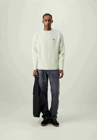 Cremfarbener gerippter Strickpullover, dunkle Jeans und schwarze Sneakers. Der Pullover hat einen runden Halsausschnitt und ein kleines Logo auf der Brust.