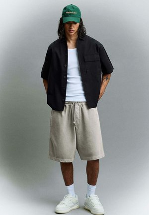 Jonge man met een groene pet met de tekst "Maybe Later", een zwart shirt met korte mouwen over een wit hemd, beige korte broek, witte sokken en witte sportschoenen.