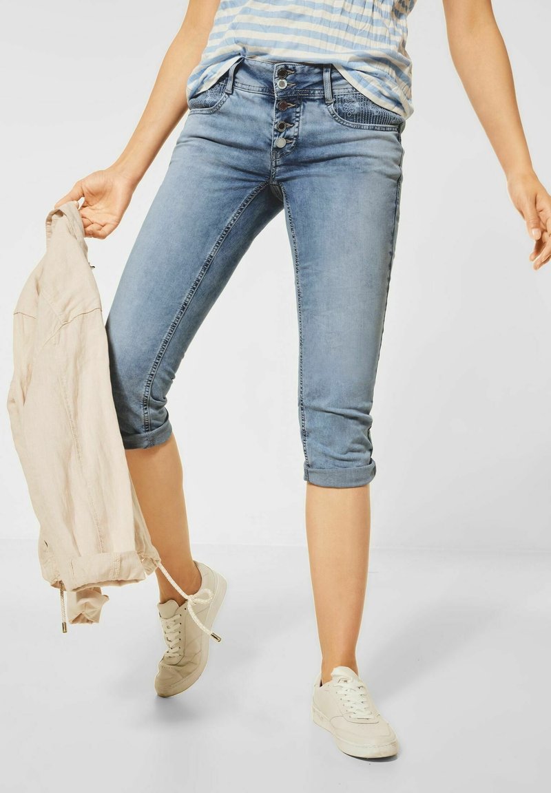 Persona che indossa pantaloni capri in denim blu arrotolati e sneakers bianche, mentre tiene una giacca beige in una mano su uno sfondo semplice.
