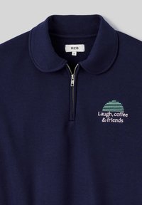Polo shirt zippé bleu marine avec un col arrondi, doté d'un design brodé multicolore comportant le texte : "Rire, café et amis." Tissu doux.
